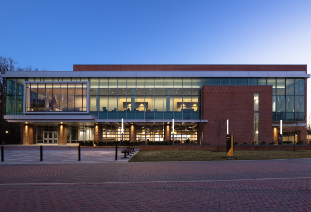 Architectural photograph illustrating the exterior of the Towson University union building. Twilight photo taken at sunset with deep blue sky, outdoor lighting, and interior lights glowing through the vast wall of large windows.